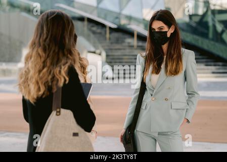 Female entrepreneurs in suits and masks standing in downtown and discussing business project while looking at each other Stock Photo