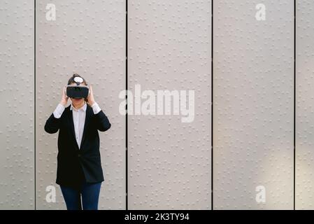 Female entrepreneur in glasses of virtual reality standing against gray wall in street and experiencing cyberspace Stock Photo