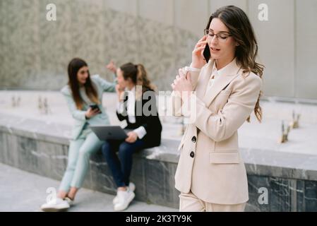 Busy female entrepreneur in formal suit standing in city center on background of colleagues and talking on mobile phone while discussing project Stock Photo