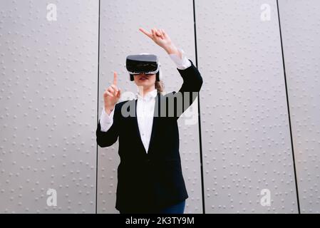 Female entrepreneur in glasses of virtual reality standing against gray wall in street and experiencing cyberspace Stock Photo