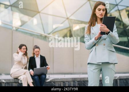 Concentrated female entrepreneur standing in downtown and browsing tablet during remote work on project on background of colleagues Stock Photo