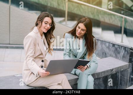 Busy female entrepreneurs in smart casual style sitting near glass office building and using gadgets while working on project together in downtown Stock Photo