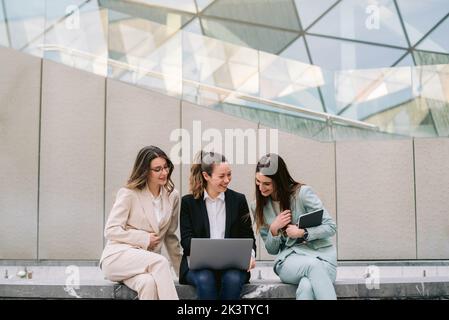 Company of smiling businesswomen in smart casual clothes sitting in city and using laptop while working on startup project together Stock Photo