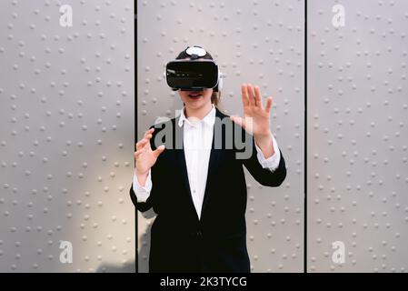 Female entrepreneur in glasses of virtual reality standing against gray wall in street and experiencing cyberspace Stock Photo