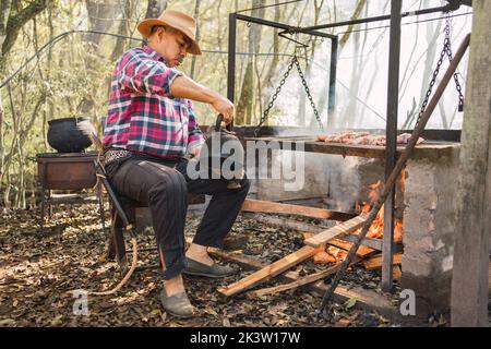 Argentinian male pouring hot water from kettle into calabash gourd against rack above burning fire in countryside Stock Photo