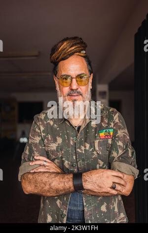 Bearded mature male in casual outfit with sunglasses and dreadlocks standing in light apartment and looking at camera Stock Photo