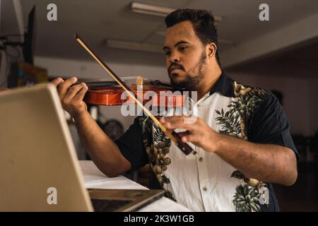 Serious young ethnic guy in casual outfit playing violin while sitting near table with netbook in light room Stock Photo