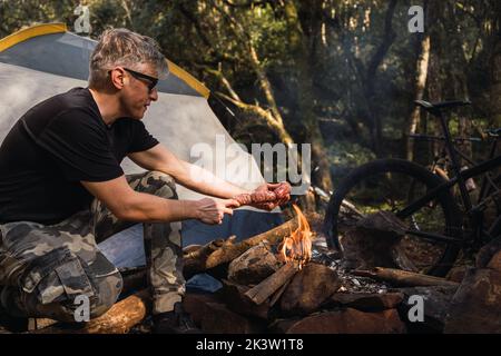 Side view of male tourist putting raw beef sausages on barbecue grill above flaming wood in campsite Stock Photo