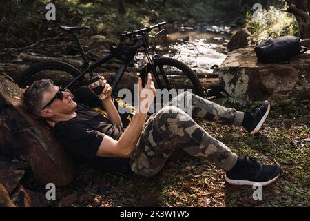 Side view of mature male traveler in sunglasses drinking hot beverage from thermos cup while sitting in campsite Stock Photo