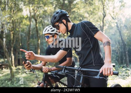 Young tattooed male bicyclist pointing with finger away showing direction while checking GPS app on cellphone during bicycle trip with friend in fores Stock Photo