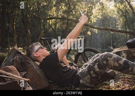 Side view of middle aged male tourist in sunglasses taking self portrait on cellphone while leaning on stone in campsite Stock Photo