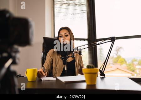 Female radio host in headphones sitting at table with microphone and taking notes in notepad Stock Photo