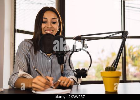Delighted female radio host sitting at table with microphone and taking notes in notepad Stock Photo