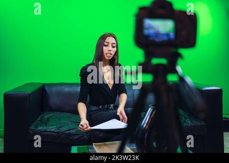 Smiling female blogger sitting on leather couch and looking at camera on tripod while recording video on chromakey background Stock Photo