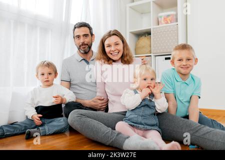 Content adult parents with charming children having fun on parquet while spending weekend in house room Stock Photo