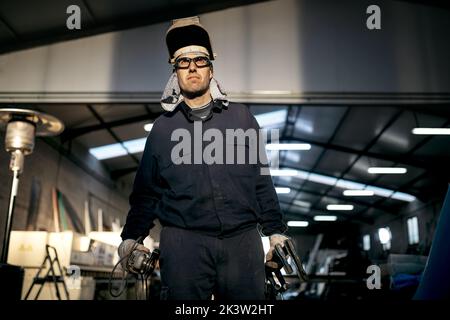 Serious workman wearing protective hardhat and blue overall while standing in garage and looking away Stock Photo