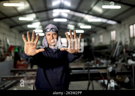 Bearded workman wearing blue overall while standing in garage and looking at camera with dirty hands Stock Photo