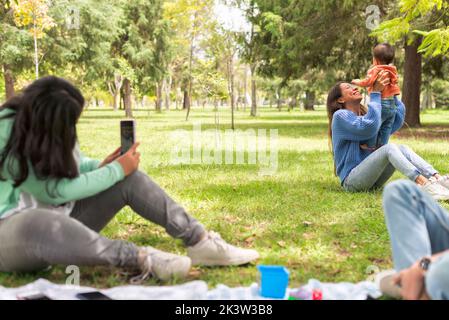 Young female taking photo on cellphone of cheerful mom raising baby while sitting on lawn in park Stock Photo