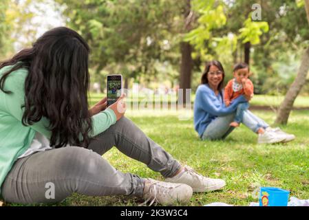 Young female taking photo on cellphone of cheerful mom raising baby while sitting on lawn in park Stock Photo