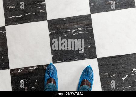 Male feet in shoe covers over classic shoes standing on stone flooring with classic checker pattern, top view Stock Photo