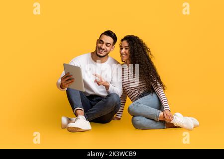 Arab Couple With Digital Tablet Relaxing On Floor Over Yellow Studio Background Stock Photo
