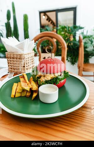 Vegan burger, in a red bun, dish with chips, on a green plate, on a wooden table, healthy vegetarian food. Stock Photo