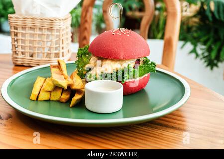 Vegan burger, in a red bun, dish with chips, on a green plate, on a wooden table, healthy vegetarian food. Stock Photo