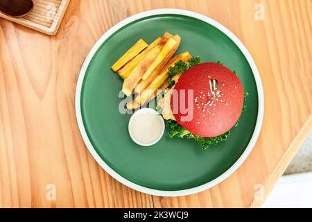 Vegan burger, in a red bun, dish with chips, on a green plate, on a wooden table, healthy vegetarian food. Stock Photo