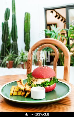 Vegan burger, in a red bun, dish with chips, on a green plate, on a wooden table, healthy vegetarian food. Stock Photo