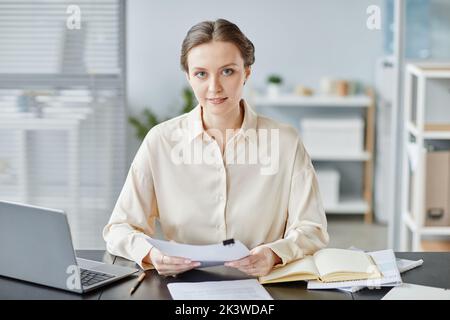 Minimal portrait of young successful businesswoman looking at camera while sitting at desk in office and holding contract Stock Photo