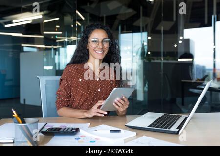 Happy and successful hispanic woman working inside modern office building, business woman using tablet computer smiling and looking at camera worker doing paperwork. Stock Photo
