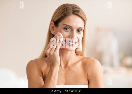 Skincare routine concept. Happy middle aged woman cleansing skin with cotton pad, standing wrapped in white towel Stock Photo