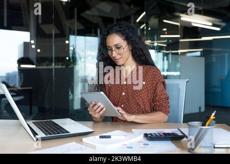 Young successful business woman working inside modern office building, Hispanic woman using tablet computer smiling and happy. Stock Photo