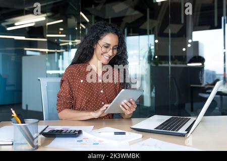 Young successful business woman working inside modern office building, Hispanic woman using tablet computer smiling and happy. Stock Photo