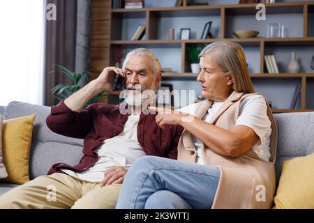 Mature couple sits anxiously at home. The gray-haired husband receives bad news on the phone. Relationships between older people Stock Photo