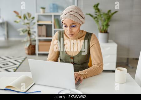 Portrait of young Middle Eastern businesswoman wearing headwrap in office and typing at laptop Stock Photo