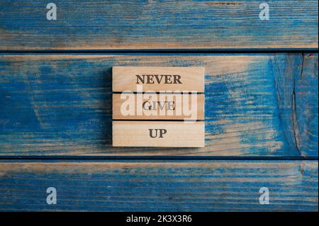 Never give up sign spelled on a stack of three woden pegs placed over textured blue wooden background. Stock Photo