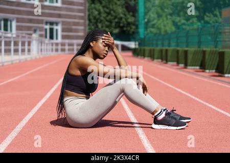 Exhausted African woman sits on track and rests after running for successful results for competition Stock Photo