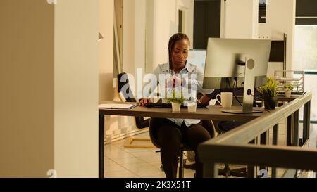 Business woman checking time on wristwatch at startup job in office with big windows, waiting on important documents. Feeling impatient to do project report with research data, take notes. Stock Photo