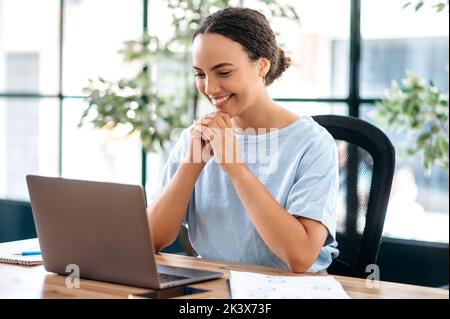 Successful confident beautiful brazilian or latino business woman, office employee, creative manager, sitting at workplace in modern office, looking at laptop, reading good news, smiling happily Stock Photo