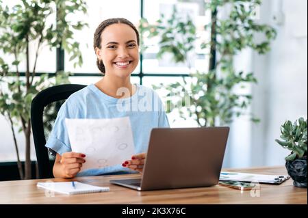 Smart successful intelligent hispanic or brazilian young woman, financial director, seo, manager, sits at a work desk in a modern office, holds documents in her hands, looks at the camera, smiles Stock Photo