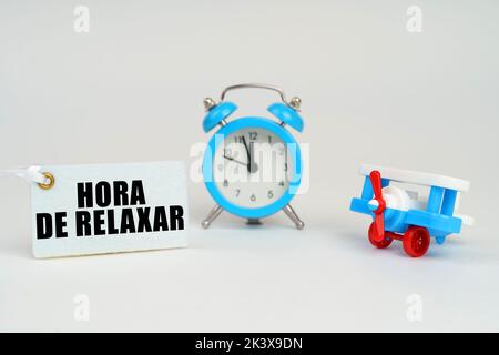 On a white surface there is a toy plane, an alarm clock and a sign with the inscription - time to relax. Business concept. Text in Portuguese. Stock Photo