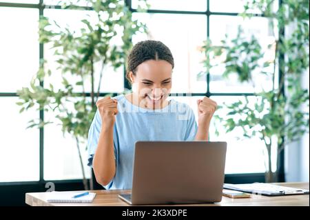 Win, success. Cheerful happy brazilian or hispanic young business woman, sits at a desk with a laptop in modern office, rejoicing in success, big profit, celebrate deal, gesturing with fists, smiling Stock Photo
