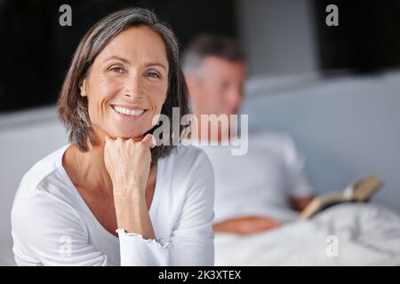 Feeling refreshed. Portrait of an attractive mature woman sitting on her bed with her husband in the background. Stock Photo
