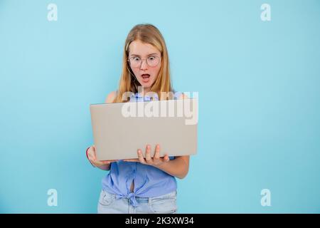 Portrait of surprised teenage girl wearing blue shirt, glasses, holding looking at screen of laptop on blue background. Stock Photo