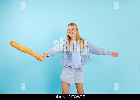 Portrait of smiling wonderful teenage girl with long fair hair wear denim clothes holding baguette on blue background. Stock Photo