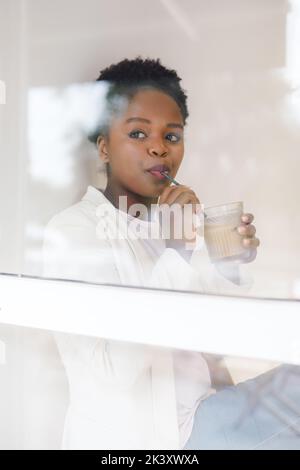 View from outside the window of an African woman enjoying a latte inside a cafe. Business owner taking a coffee break while working in the office. Stock Photo