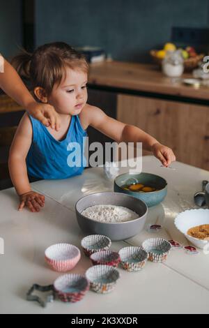 A mother and her baby daughter cooking in kitchen, on a wooden table full of ingredients and a baking forms. Preparing pancakes for breakfast at home Stock Photo