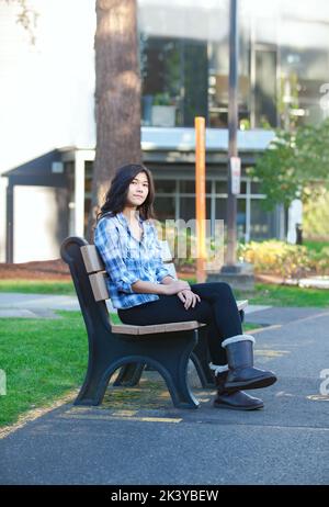 Biracial Asian teen girl sitting on bench outside school building, legs crossed Stock Photo