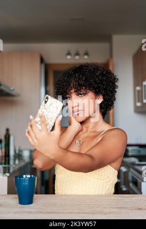 Confident young transgender woman taking selfies with a smartphone while sitting in the kitchen at home. Stock Photo